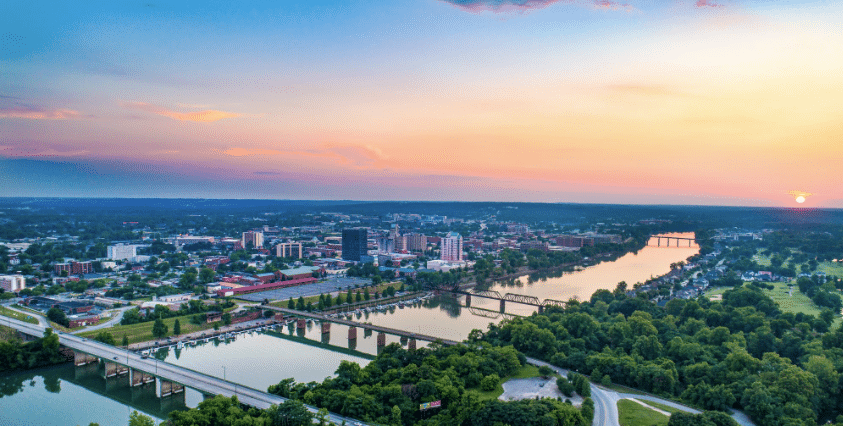 Panoramic view of Augusta, Georgia, at sunset, with the Savannah River in the foreground and the city skyline against a pastel-colored sky. The sun is visible near the horizon, casting a warm glow over the landscape.