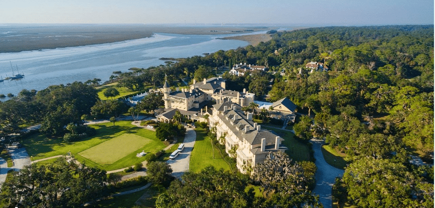 Aerial view of Jekyll Island featuring a large, luxurious estate with expansive manicured lawns adjacent to a vast river. The property is surrounded by lush greenery, with a forest canopy extending to the horizon, and a clear sky above. Several smaller buildings and a boat are also visible along the water's edge, emphasizing the island's serene coastal environment.