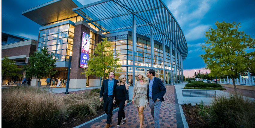 Group of four adults walking and chatting near the modern glass facade of the Sandy Springs Performing Arts Center in Sandy Springs, GA, during dusk.