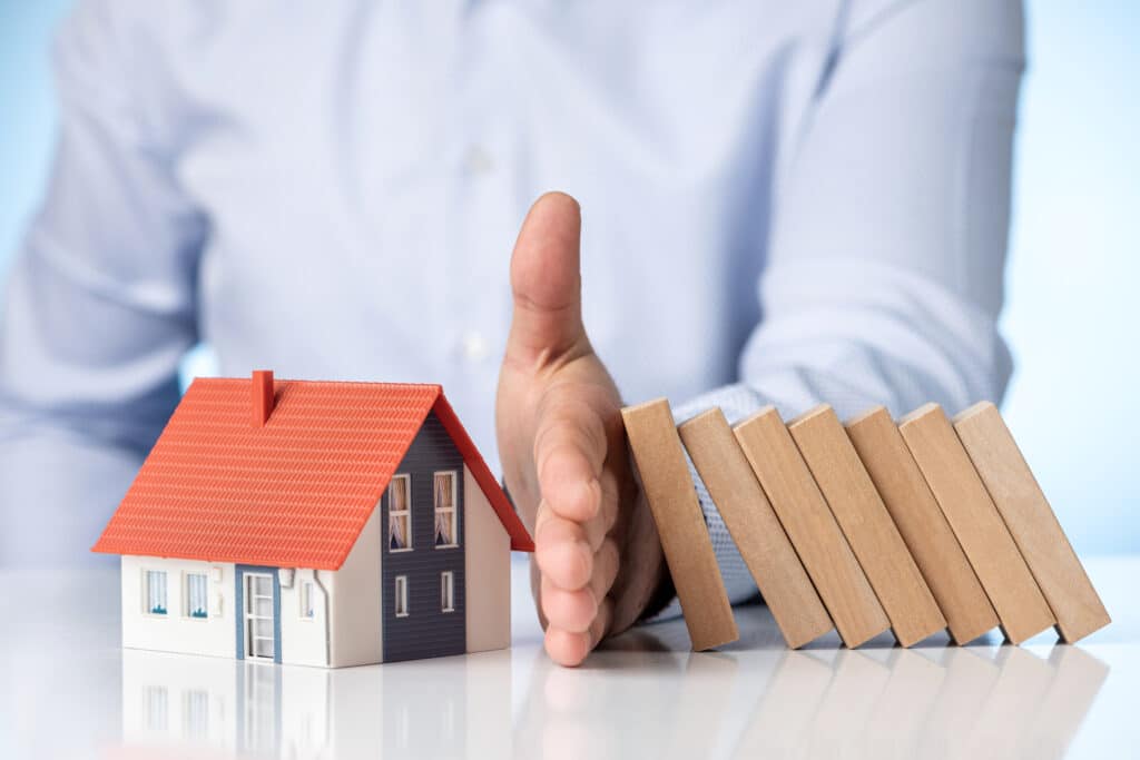 a hand stopping a row of falling dominos from hitting a model house, symbolizing protection or prevention. This is often used as a metaphor for safeguarding a home from potential risks or disasters, representing the concept of home insurance and risk management.