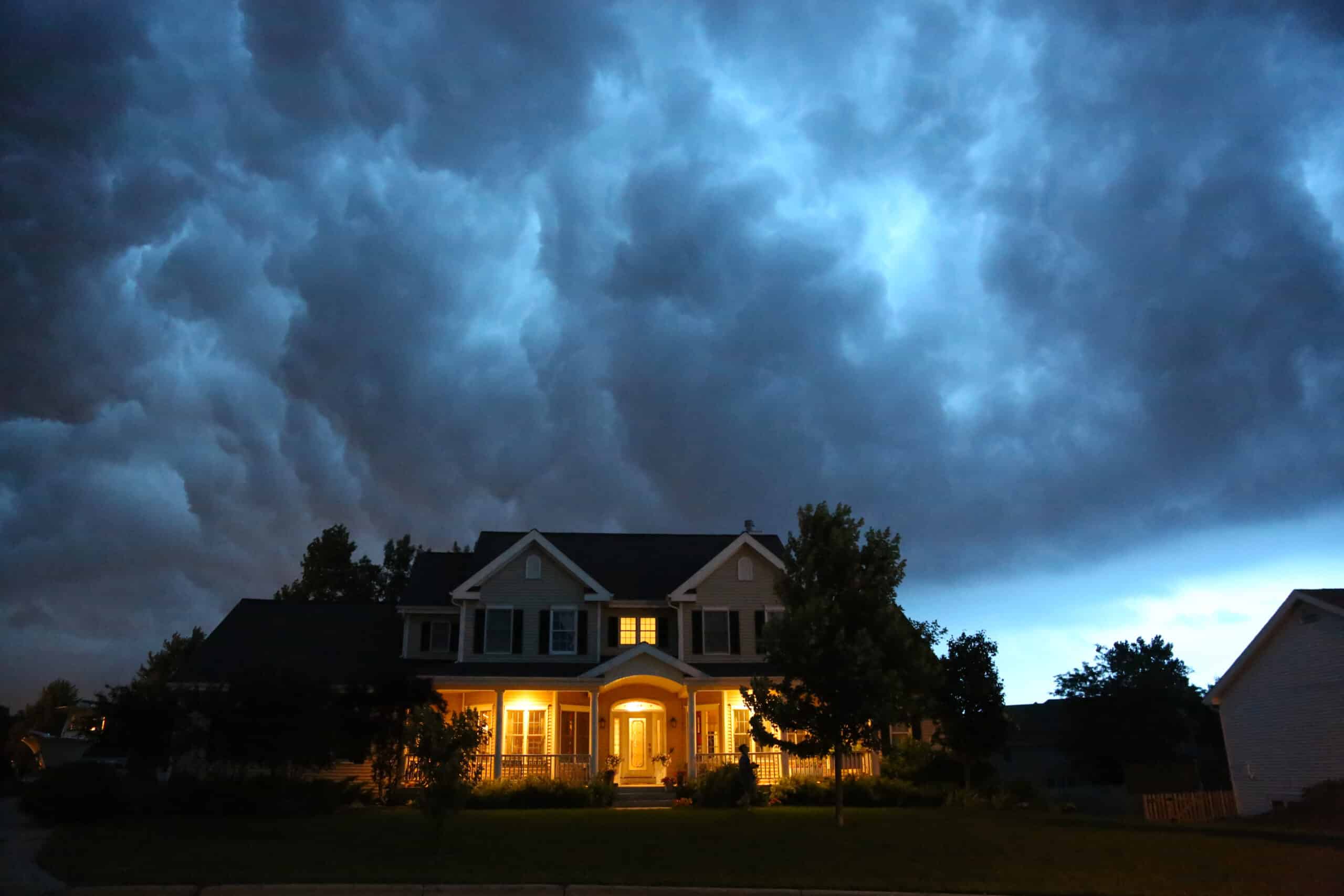 A two-story residential house during twilight or early night, with lights turned on inside and around the porch area. The sky above is dramatic and tumultuous with dark storm clouds, suggesting an impending or ongoing storm. The contrast between the warm lights of the house and the dark, foreboding sky adds a sense of coziness to the home, while simultaneously creating an atmosphere of tension due to the storm. This scene could be used to illustrate concepts such as the safety and shelter a home provides against the elements, the onset of severe weather, or the importance of home insurance in protecting against natural disasters.