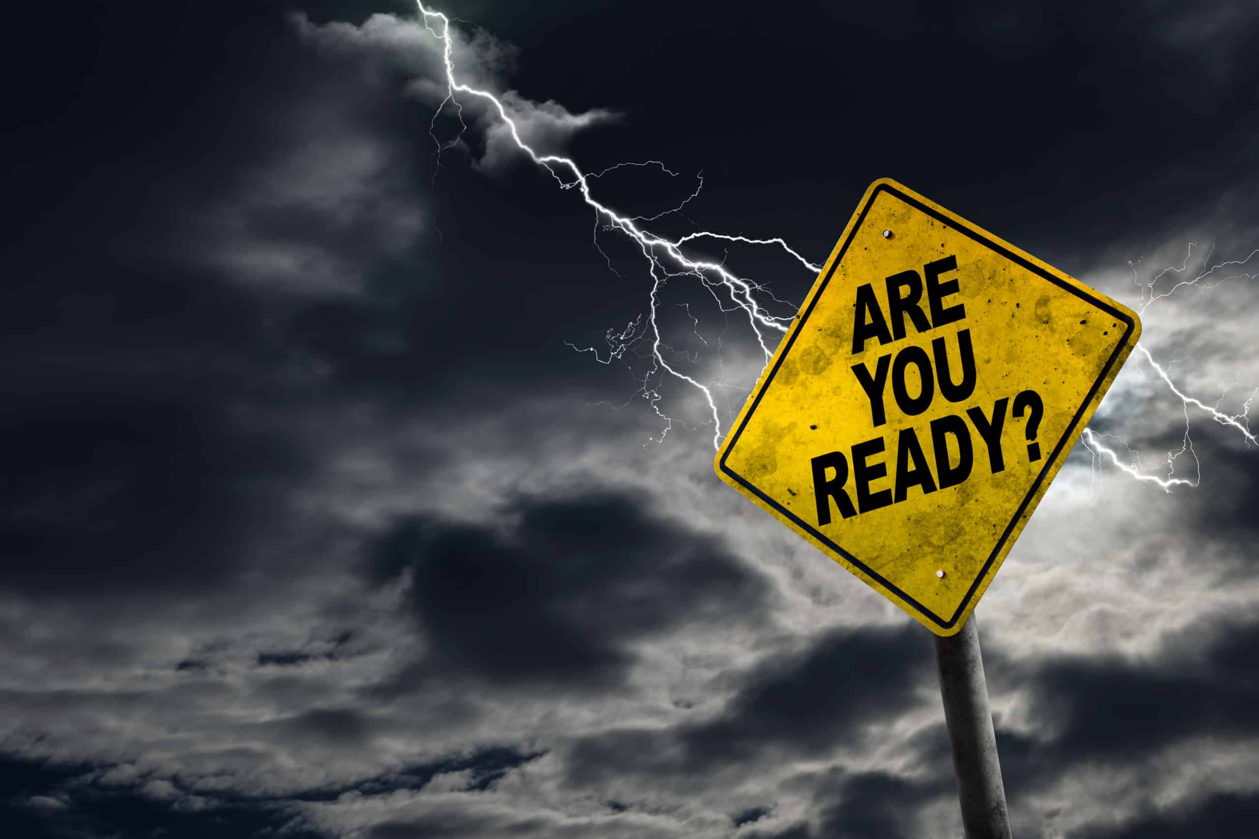 The image shows a weathered yellow road sign with the words "ARE YOU READY?" in bold, black letters. The sign stands against a dramatic backdrop of a stormy sky with dark clouds and multiple branches of lightning striking down. The scene suggests a sense of urgency or impending challenge, perhaps as a metaphor for preparation and resilience in the face of difficult circumstances.