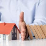 A scene often used to represent the concept of protection or insurance in a metaphorical way. There's a hand of a person, appearing to stop a row of falling dominos from hitting a small model house with a red roof. This setup typically symbolizes safeguarding a home from potential harm or financial distress.