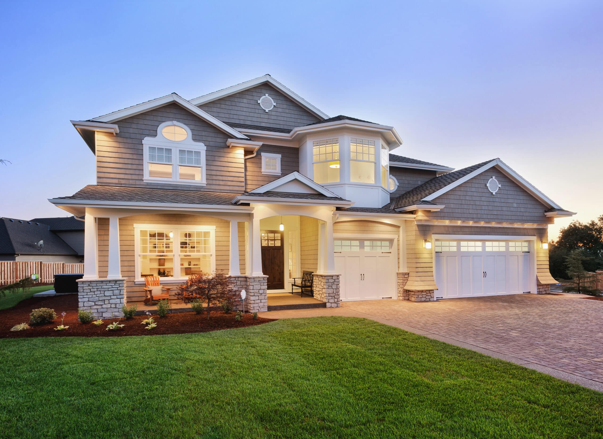 a large, two-story house during twilight. The house features a mix of craftsman and modern architectural styles, with a facade combining stone accents at the base of the columns and grey siding. It has a prominent front porch lit warmly, invitingly with chairs visible, suggesting a cozy seating area. The roof is shingled, with several gables adding to the architectural interest. A three-car garage is visible to the right, with white doors that match the trim around the windows and doors of the house. The landscaping is well-maintained with a manicured lawn, shrubs, and a few small trees, leading up to a stone pathway to the entrance. The overall impression is of a spacious, inviting home in a suburban setting, captured at a moment when the lighting enhances its welcoming ambiance.