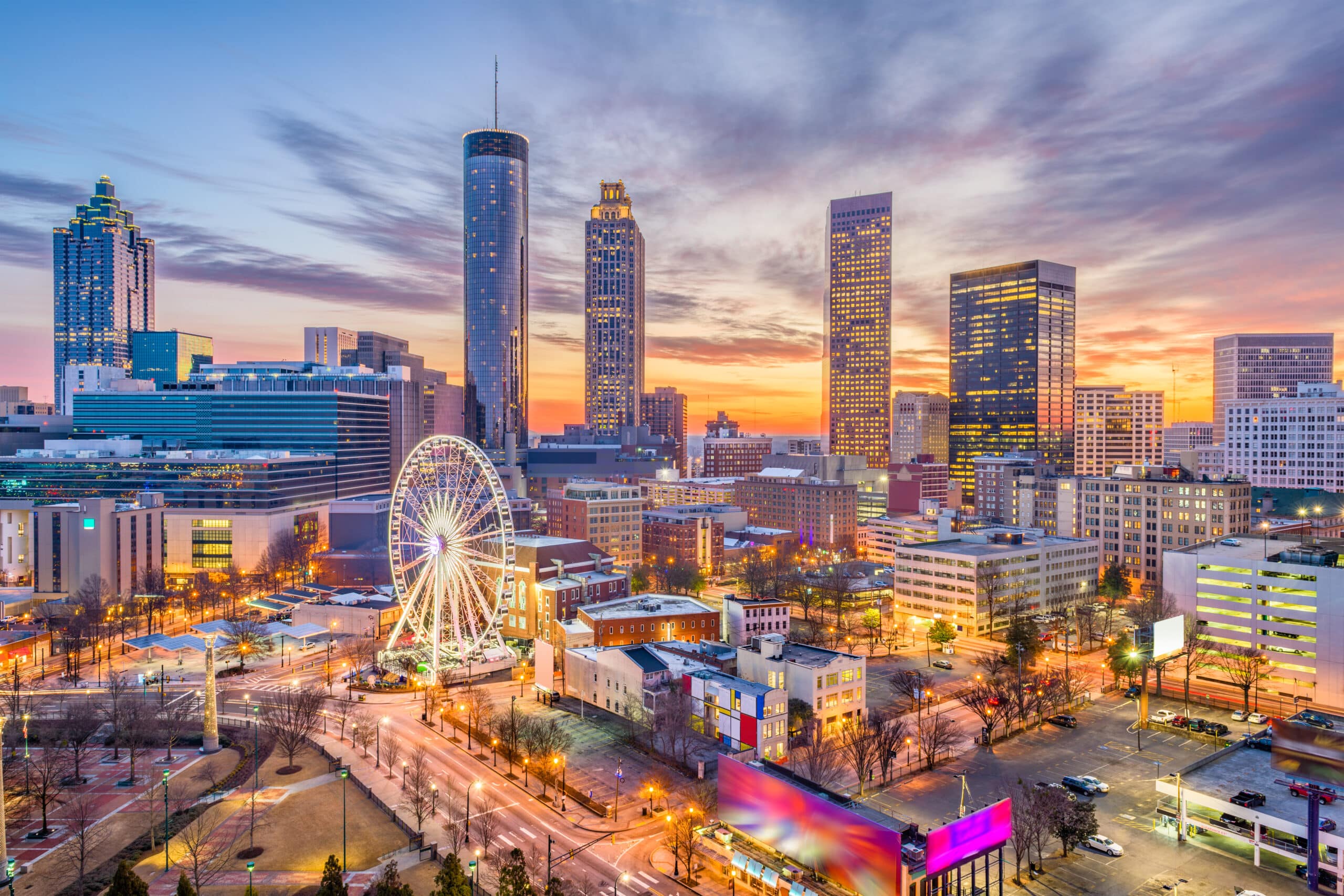 A panoramic view of Atlanta Georgia at dusk, featuring modern skyscrapers, a lit Ferris wheel, and historic buildings under a colorful sunset sky.