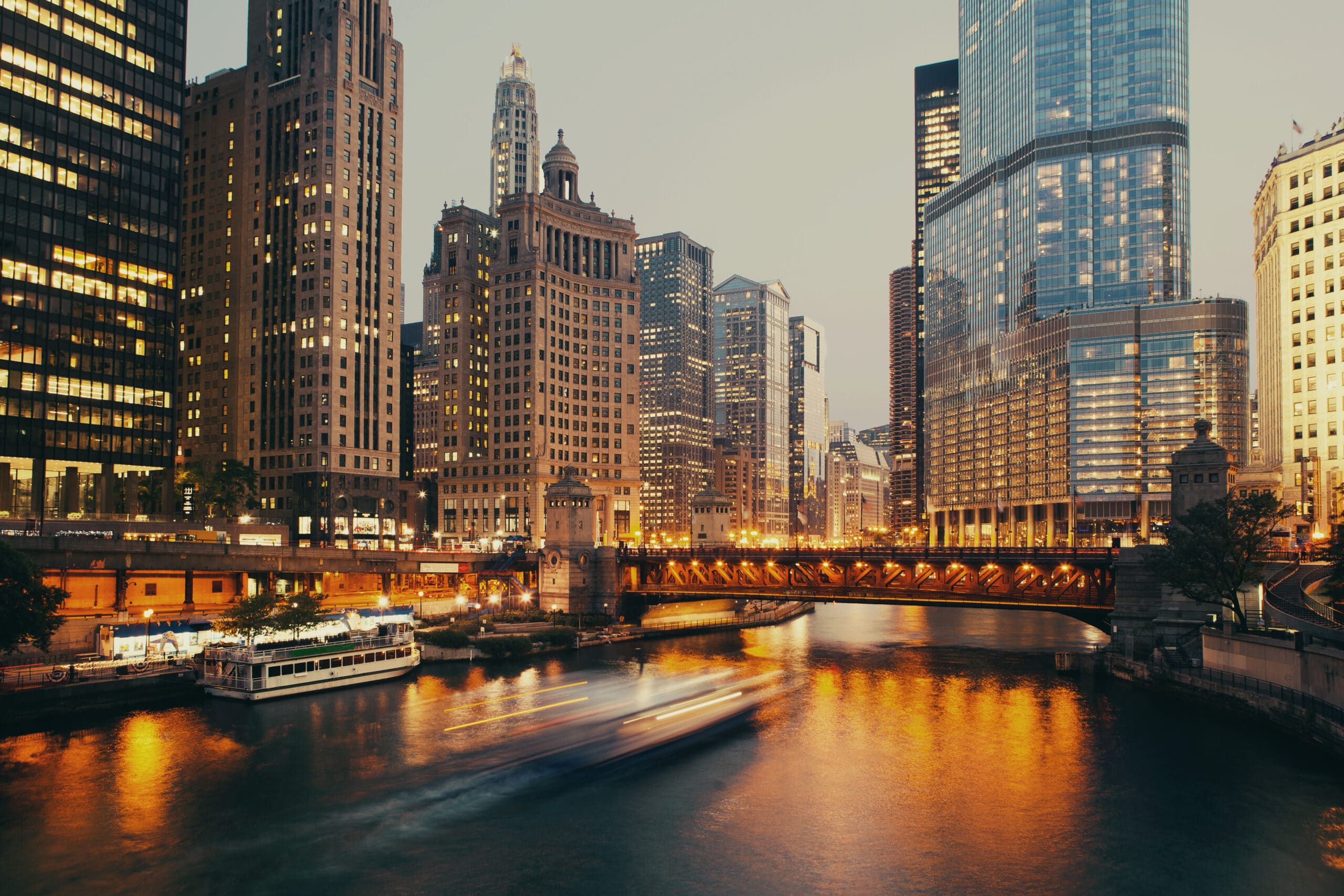 Chicago at dusk with illuminated buildings along a river. The architecture blends historical and modern styles, with skyscrapers and a bridge visible. The scene is warm and inviting, showcasing the vibrant city life and peaceful water reflections.