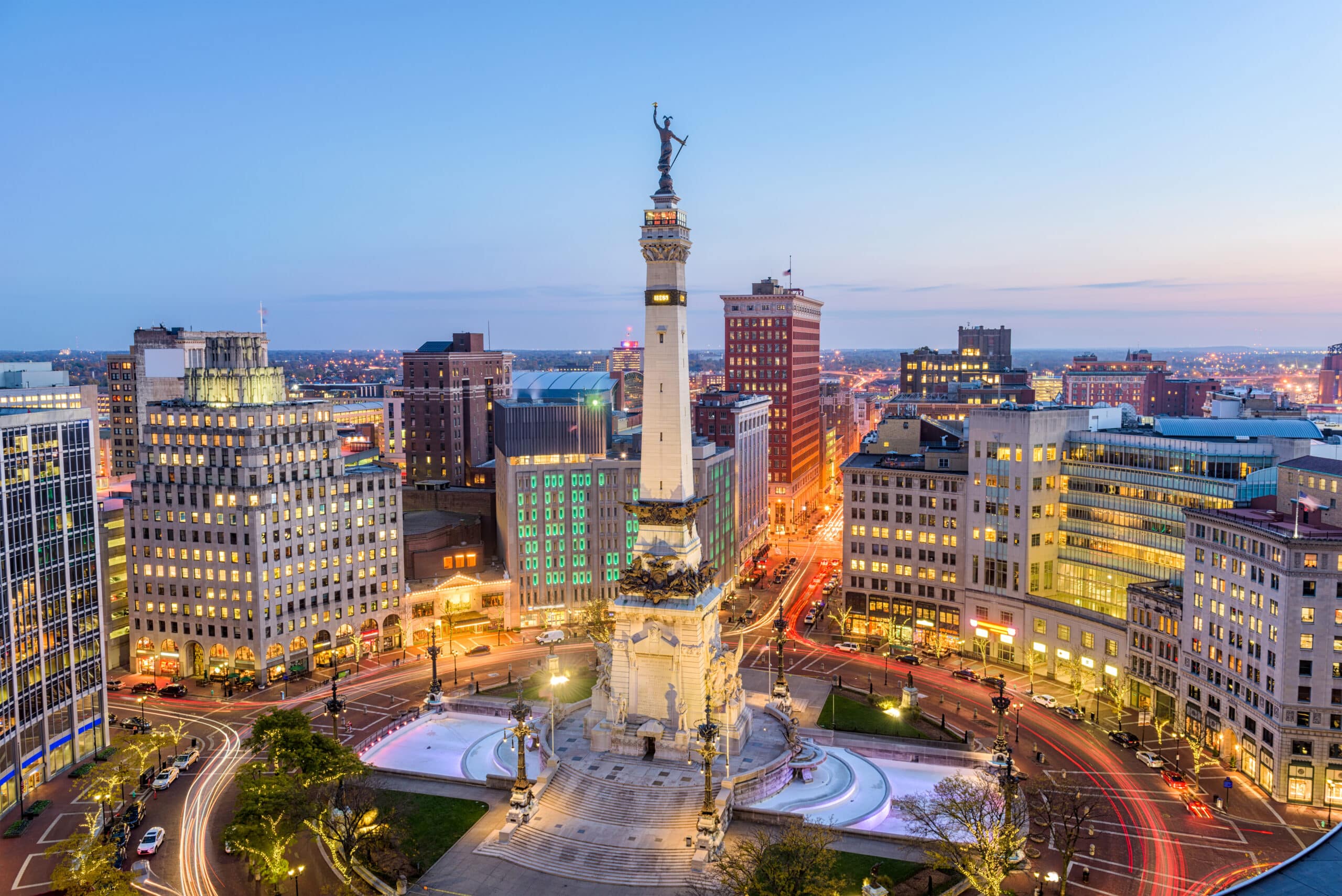 Twilight view of a bustling cityscape with a prominent monument at the center, surrounded by circular patterns on the ground, illuminated by streetlights and building lights, with a mix of historic and modern buildings and light trails from moving vehicles, against a transitioning sky from sunset to night.