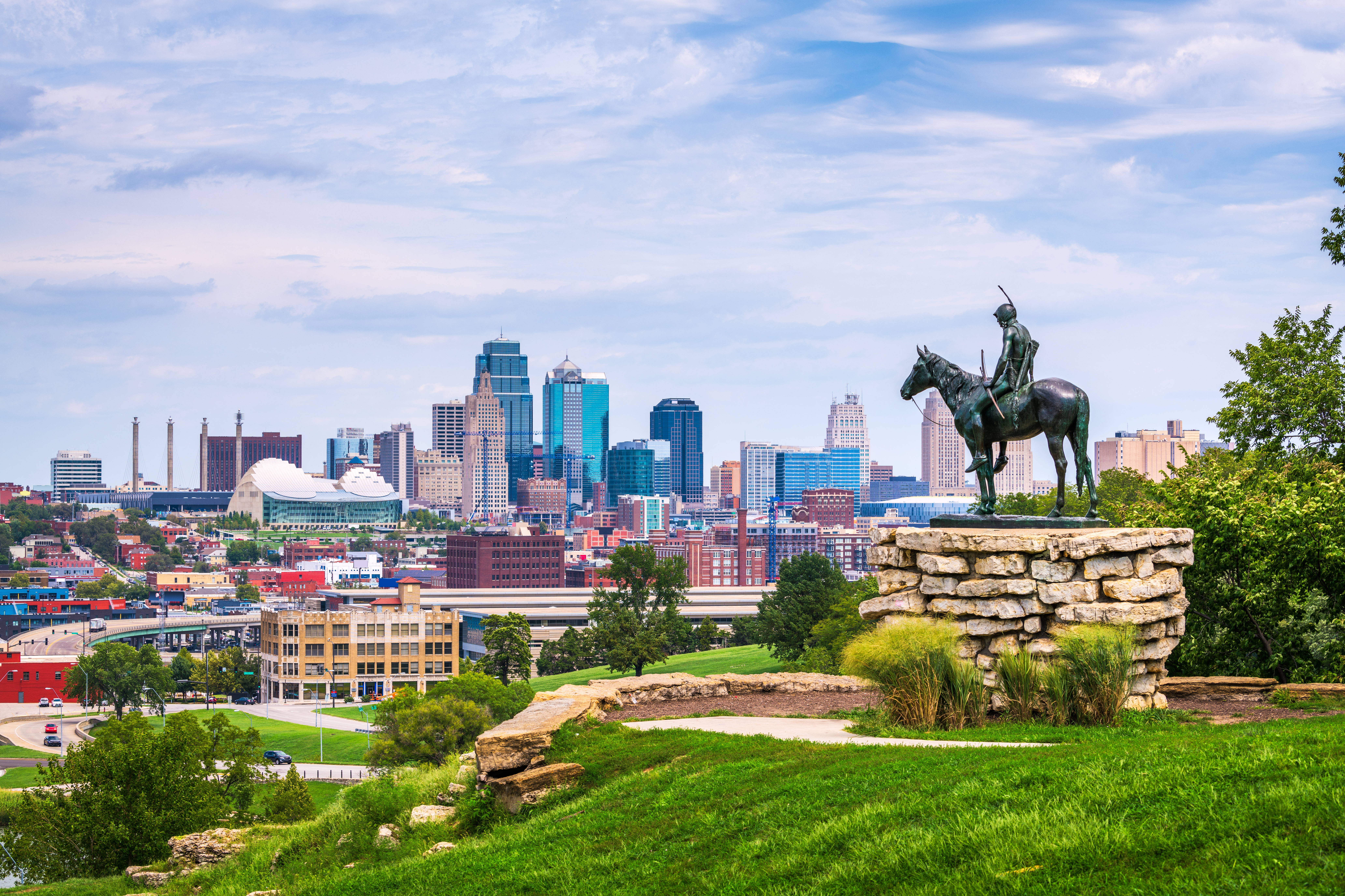 A vibrant city skyline with various skyscrapers in the background, viewed from a vantage point featuring a statue of an individual on horseback in the foreground, set upon a stone pedestal surrounded by lush greenery.