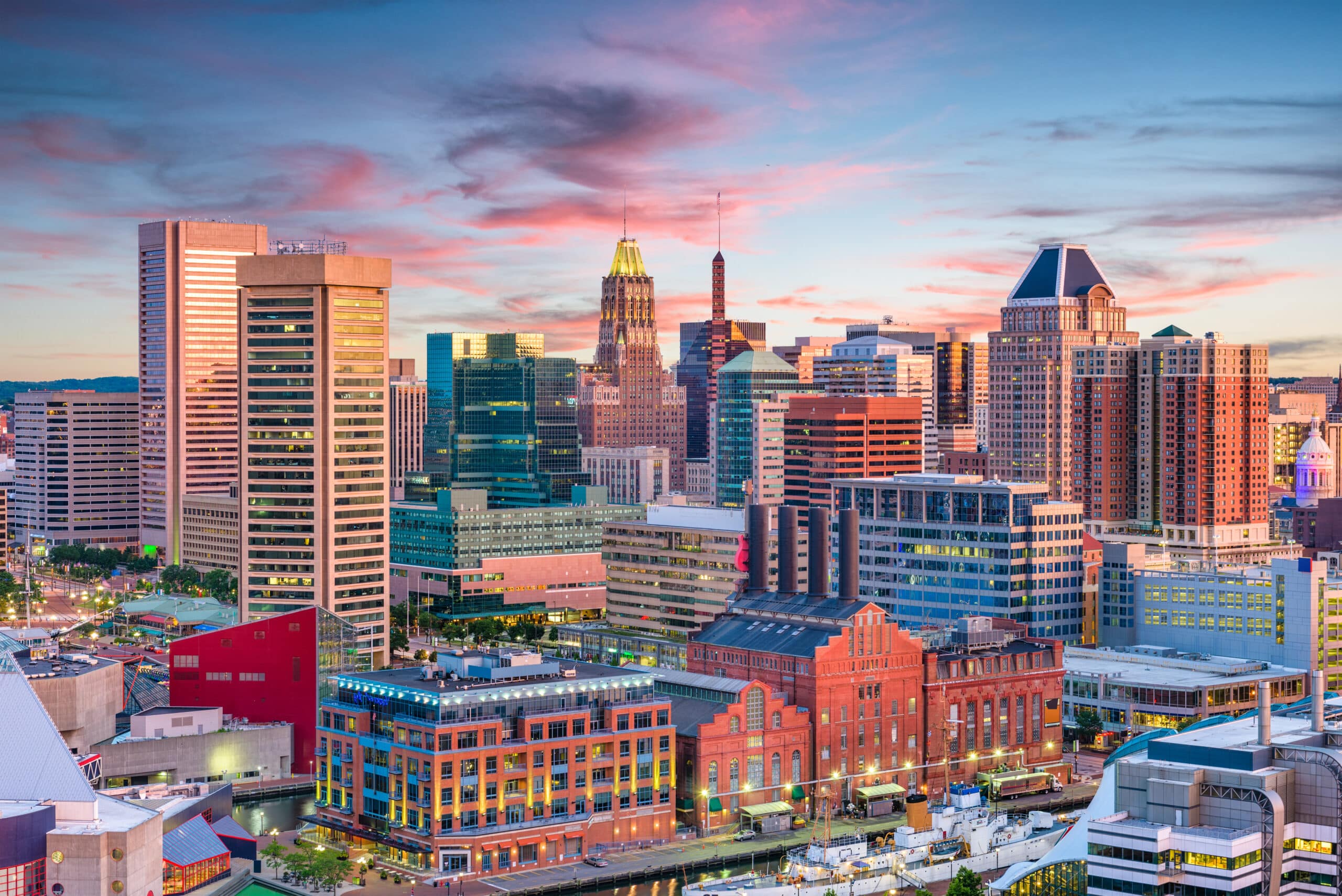 A vibrant city skyline at sunset, featuring a mix of historic and modern buildings. The setting sun casts a warm glow, highlighting the architectural diversity against a backdrop of pink, orange, and blue sky. Skyscrapers, commercial spaces, and residential areas are visible, reflecting the city's dynamic atmosphere.