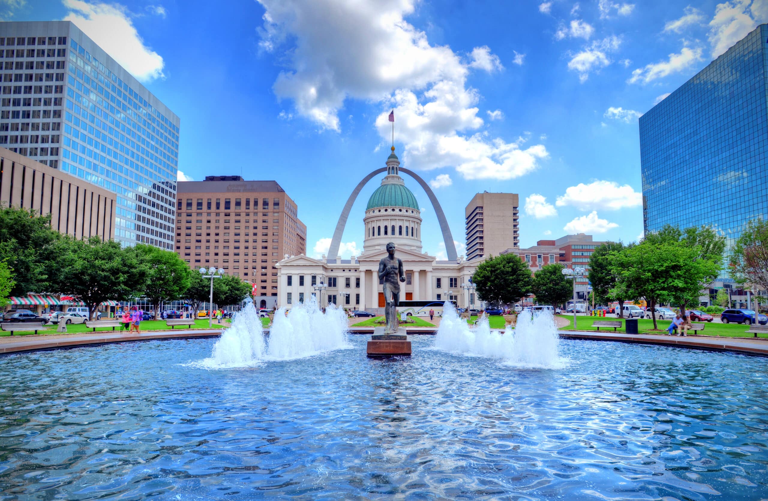 A vibrant city park with a fountain in the foreground and the iconic Gateway Arch in the background, under a clear blue sky with scattered clouds. The scene includes a statue, green spaces, and surrounding buildings of varying architectural styles.