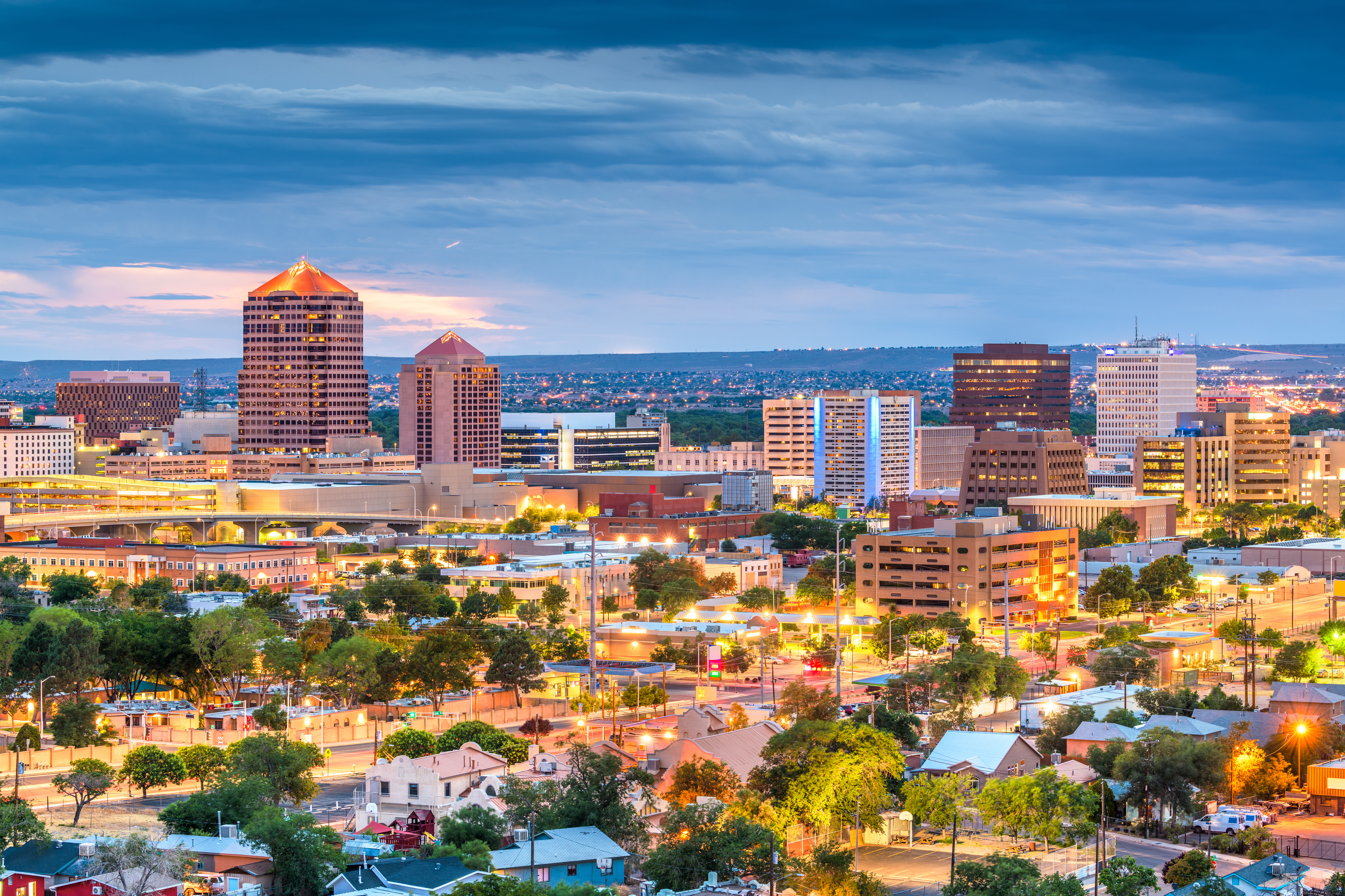 A cityscape during twilight, showcasing a blend of modern and traditional architecture. Prominent are two pyramid-shaped buildings that stand out against the skyline, illuminated by the soft glow of sunset. The city is densely populated with various structures, including residential buildings, office towers, and possibly commercial spaces. The foreground features residential areas with greenery interspersed, leading up to the more densely built-up city center. The lighting suggests early evening, with street lights beginning to glow and the interiors of buildings illuminated. The backdrop includes a vast, open sky with hues of blue and pink, indicating the transition from day to night. Overall, the photo captures the dynamic nature of an urban environment at dusk, highlighting architectural diversity and the bustling activity of city life.