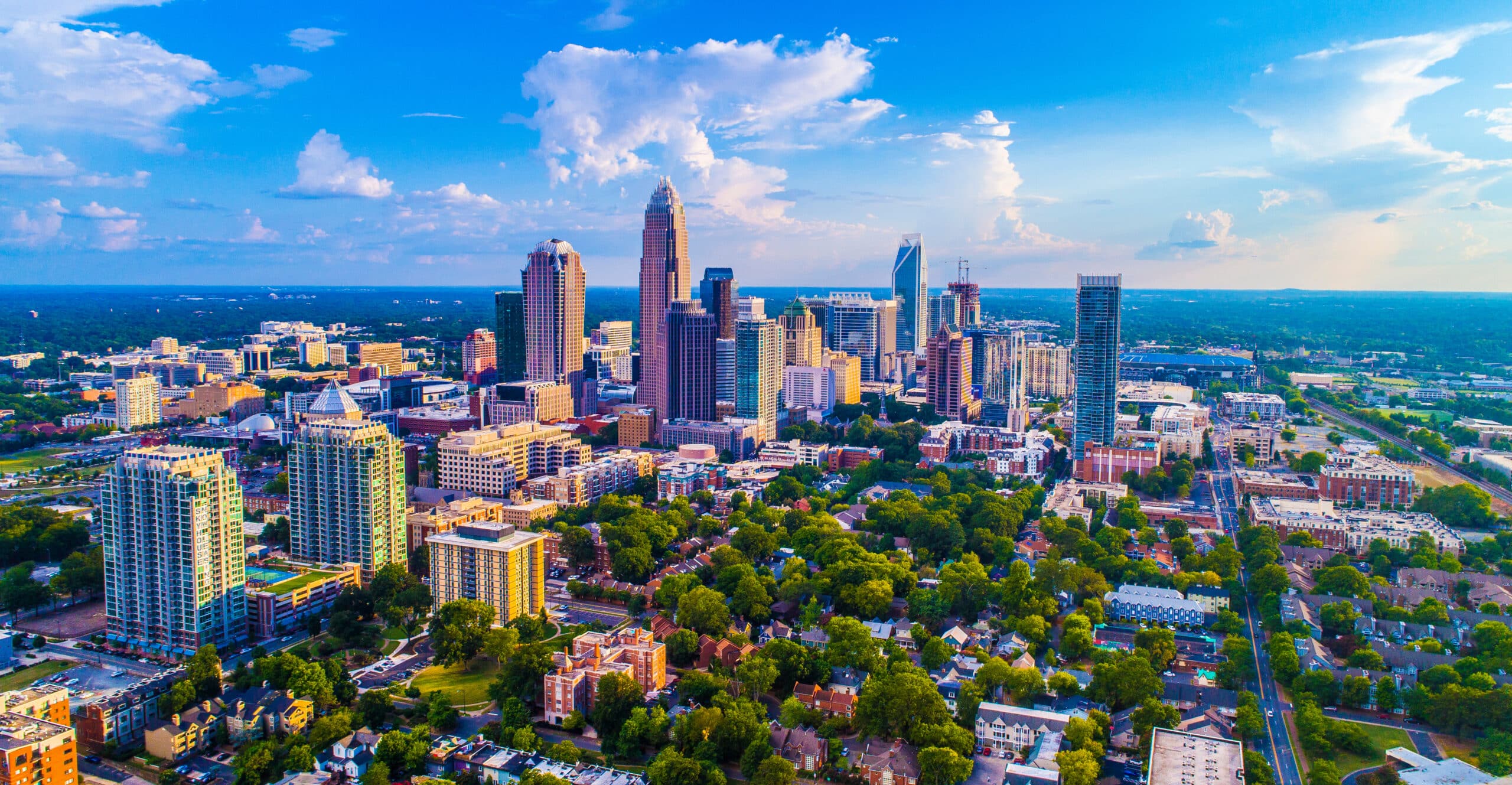 Aerial view of the densely populated city of Charlotte with a mix of skyscrapers and residential buildings, surrounded by greenery under a blue sky with scattered clouds.