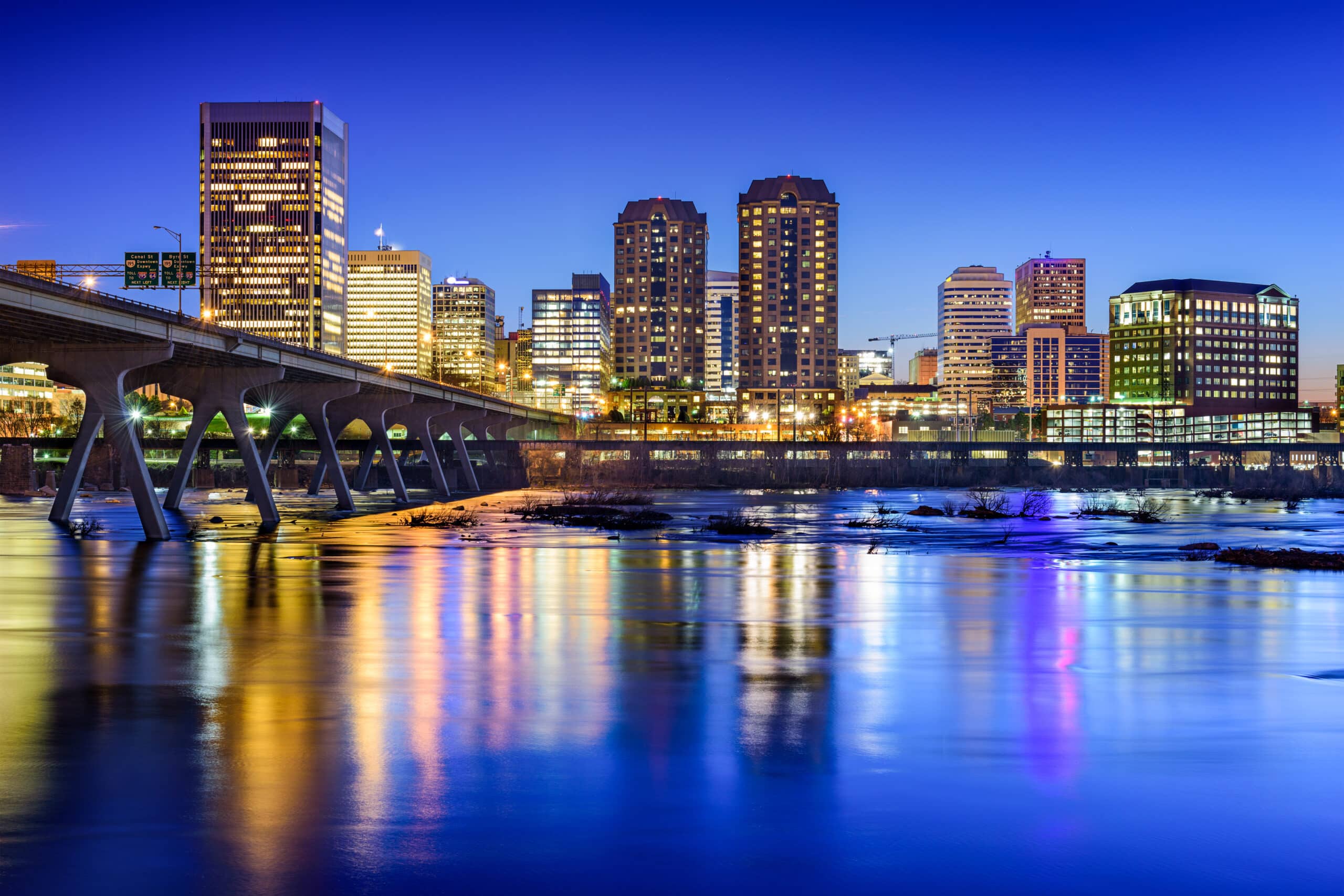 Richmond, Virginia skyline at twilight with illuminated buildings reflecting in the calm river below. A bridge with distinctive supports crosses the river, leading into the bustling downtown area. The scene combines natural and urban elements, showcasing a harmonious transition from day to night.