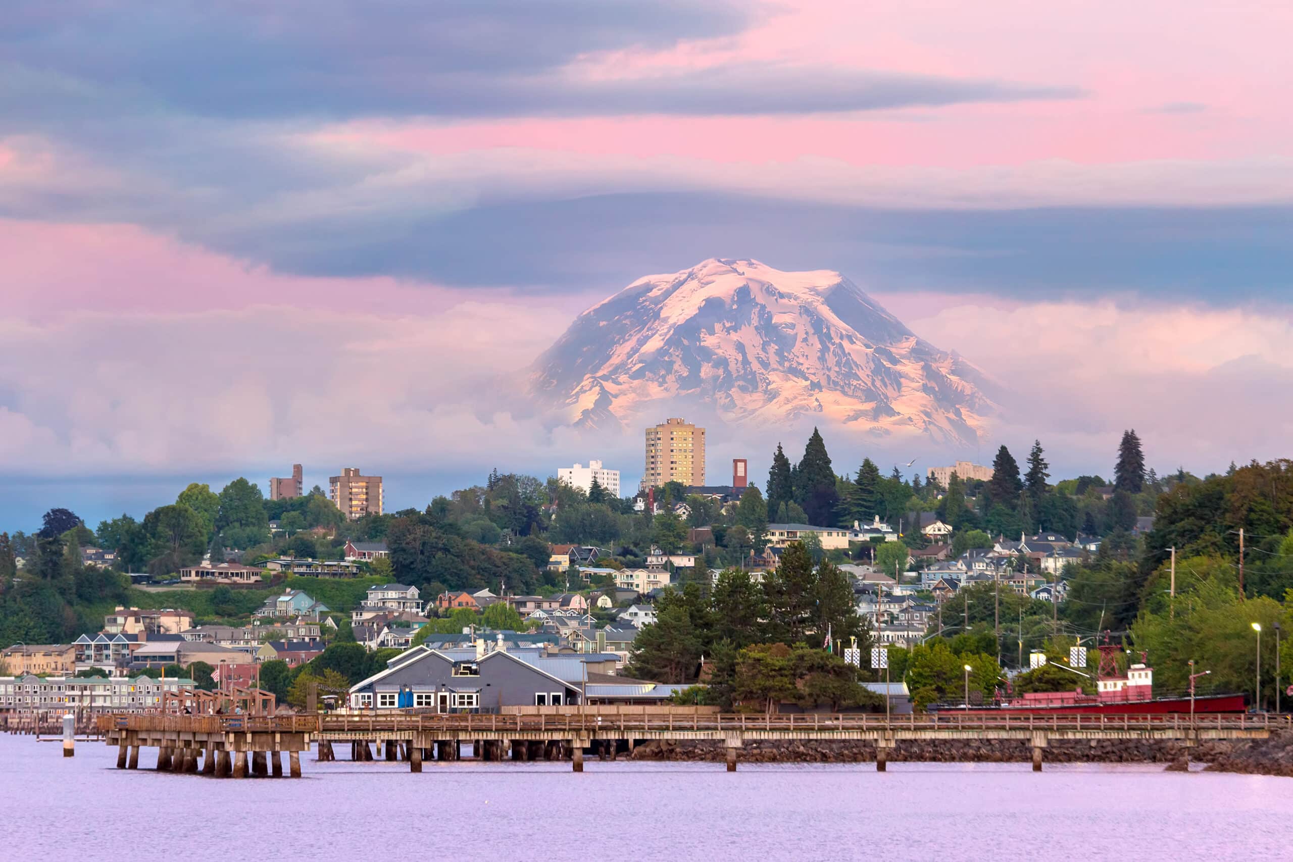 A picturesque view of a waterfront city at sunset with a snow-capped mountain in the background, under a soft pink sky