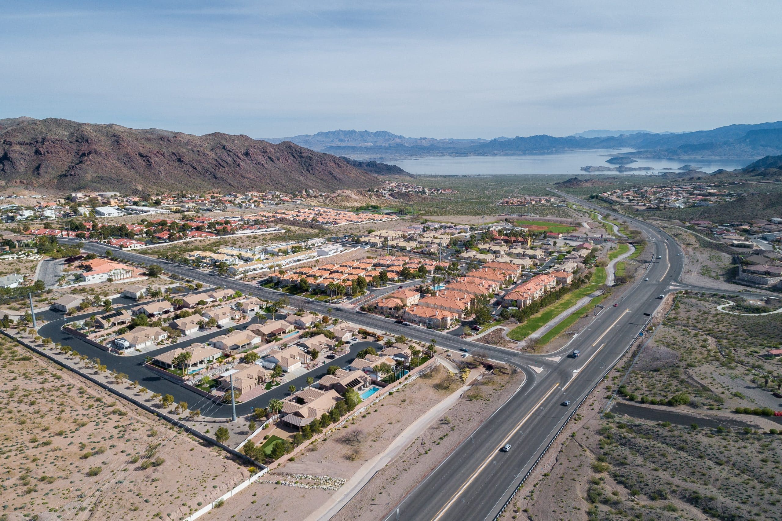 The Boulder City, NV area is nestled between a mountain range and a large body of water, which could be a lake or a reservoir. The layout shows a major roadway cutting through the town, lined with a mix of residential neighborhoods featuring predominantly orange-tiled roofs, typical of arid climate regions. The image captures the contrasting natural and developed land, offering a clear view of how the community is organized around the natural geography.