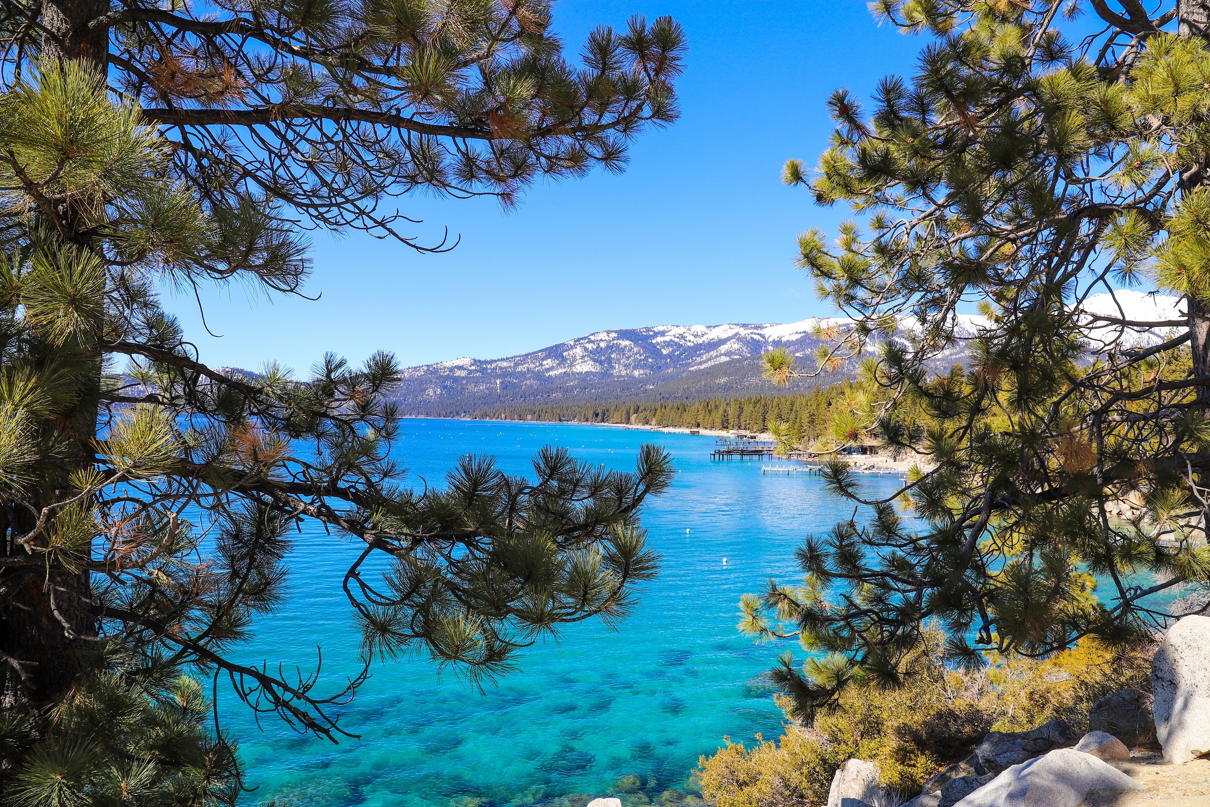 a beautiful and serene image of a lake with clear turquoise waters, framed by lush pine trees. In the background, there are snow-capped mountains, and you can also see some structures that appear to be docks or small piers along the lakeshore. The sky is clear and blue, contributing to a vibrant and picturesque scene. This place seems perfect for a peaceful getaway or a nature retreat.