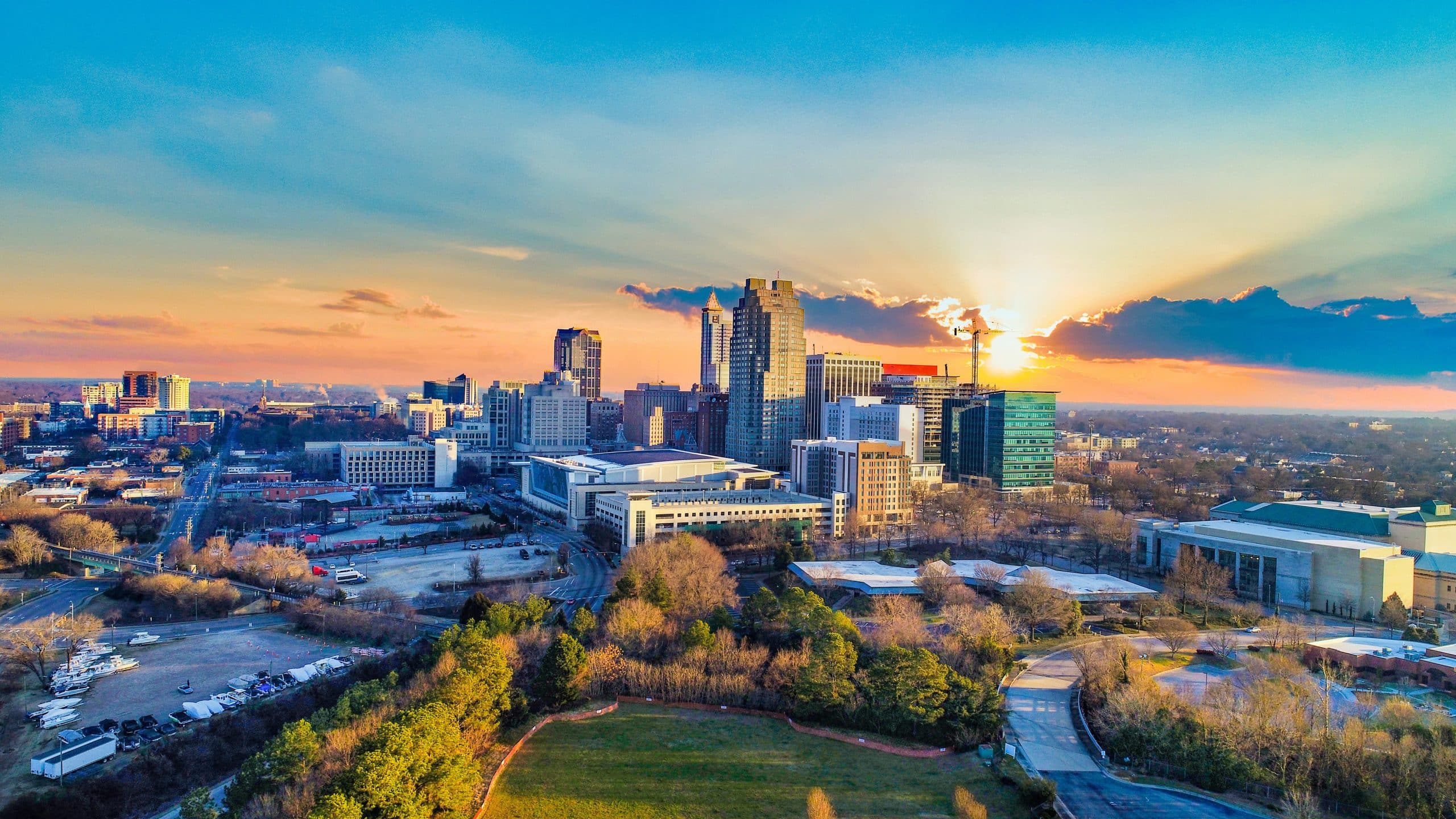 Aerial view of Raleigh, North Carolina at sunset. The city skyline features modern buildings illuminated by the setting sun, which casts a warm glow over the horizon. Below the skyline, there are green spaces and a mix of residential and commercial areas. The sky is a blend of orange and blue hues with some scattered clouds, enhancing the picturesque scene.