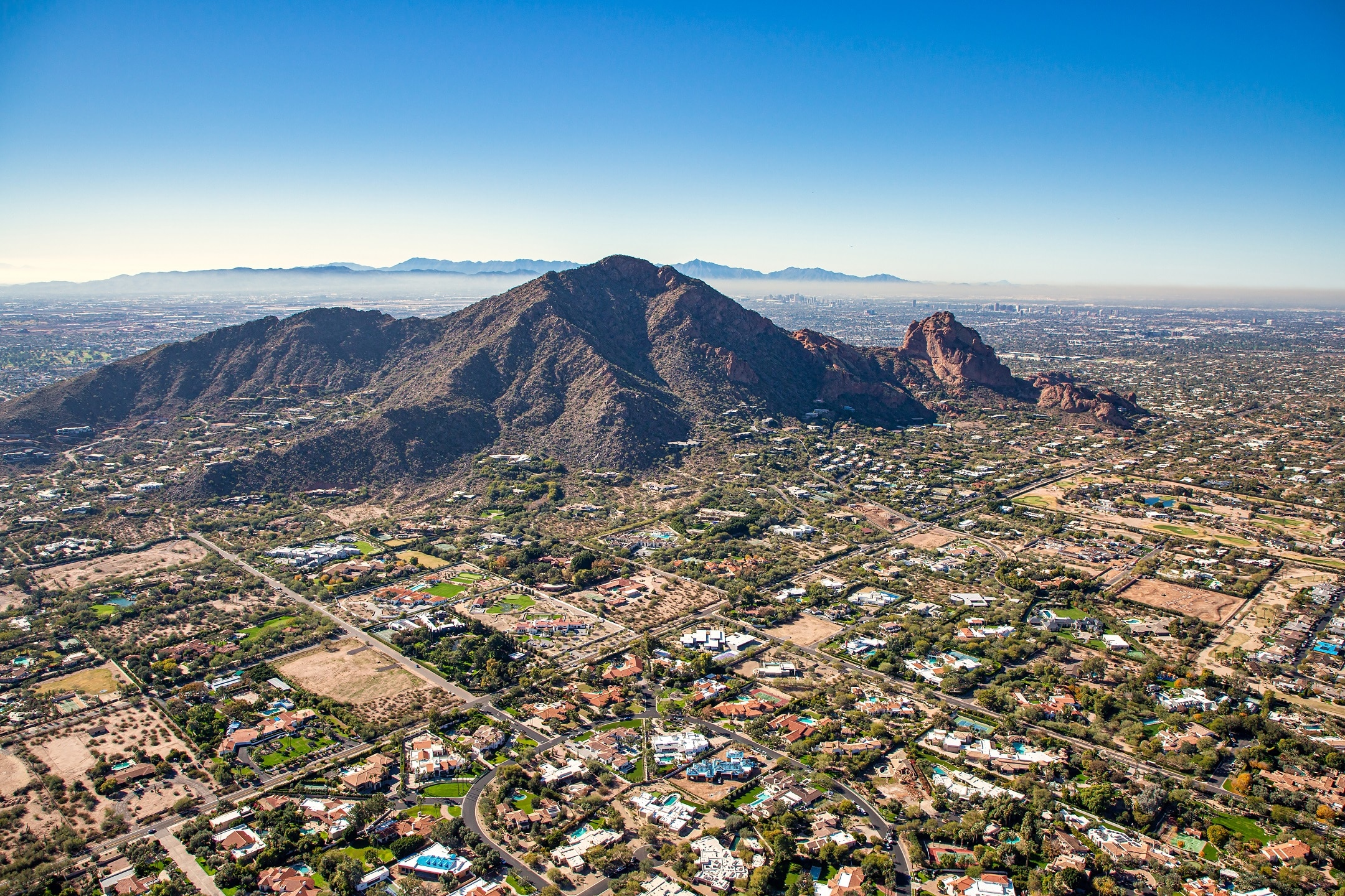 A stunning aerial view of a landscape in Paradise Valley AZ with a significant contrast between natural and urban elements. Dominating the scene are rugged, mountainous terrains with their peaks creating a dramatic backdrop. The foreground is filled with a sprawling urban area, characterized by numerous residential and commercial buildings interspersed with green spaces, which suggests a well-integrated city layout. The city extends into the plains, fading into the hazy horizon where other mountain silhouettes can be discerned. This vivid image effectively showcases the intersection of nature and human habitation, highlighting both the beauty of the natural environment and the density of the urban settlement.