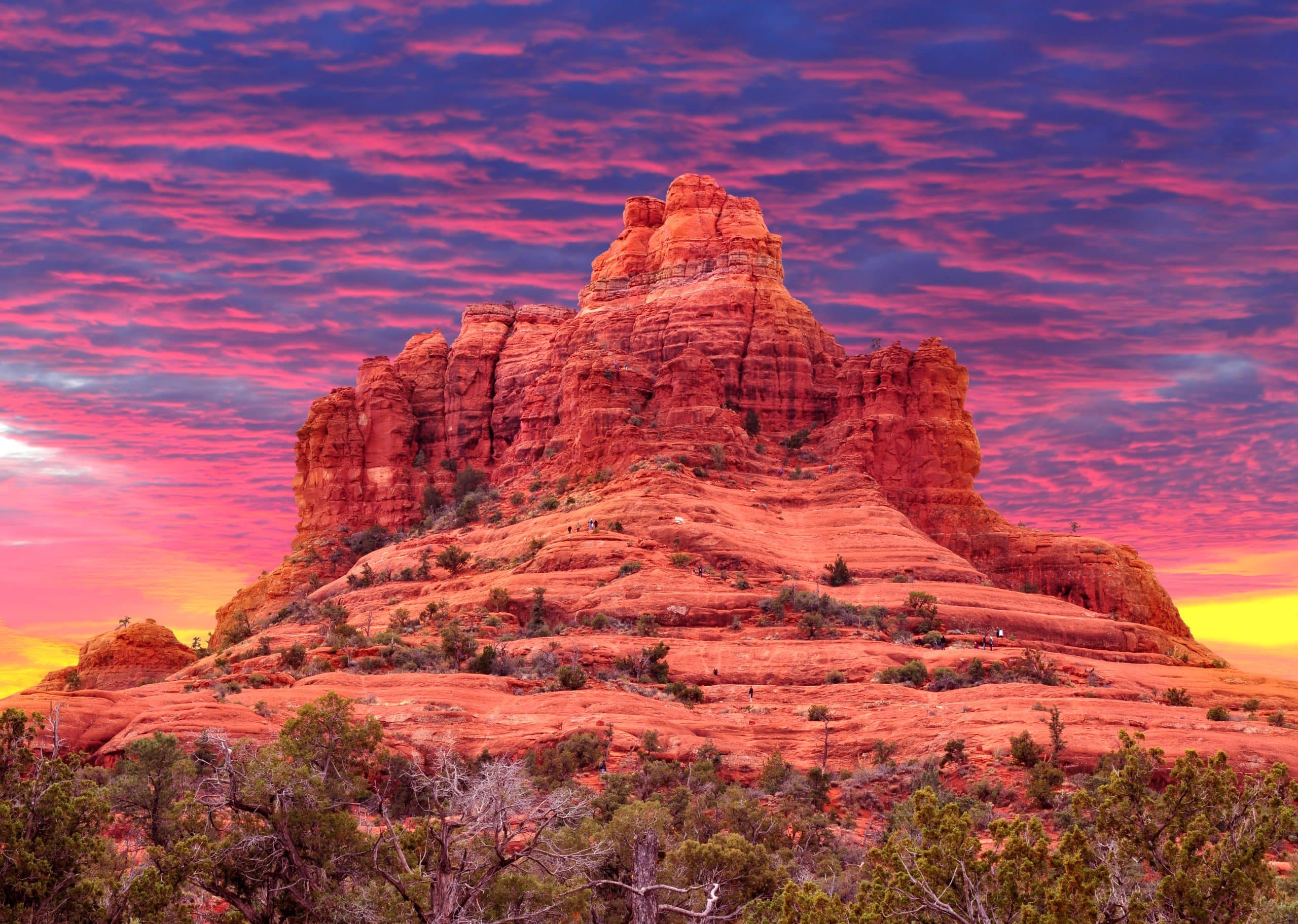 The image features a strikingly vibrant landscape at sunset with a dramatic rock formation. The sky is filled with a canvas of rich, deep pinks and purples, blending into fiery orange and yellow at the horizon. The rock formation, composed of layered red sandstone, dominates the foreground, standing tall against the colorful sky. This geological feature is weathered and rugged, characterized by its unique contours and peaks. Sparse vegetation and a few small trees dot the red, undulating terrain around the base of the rock, adding a touch of green to the predominantly red and orange scene.