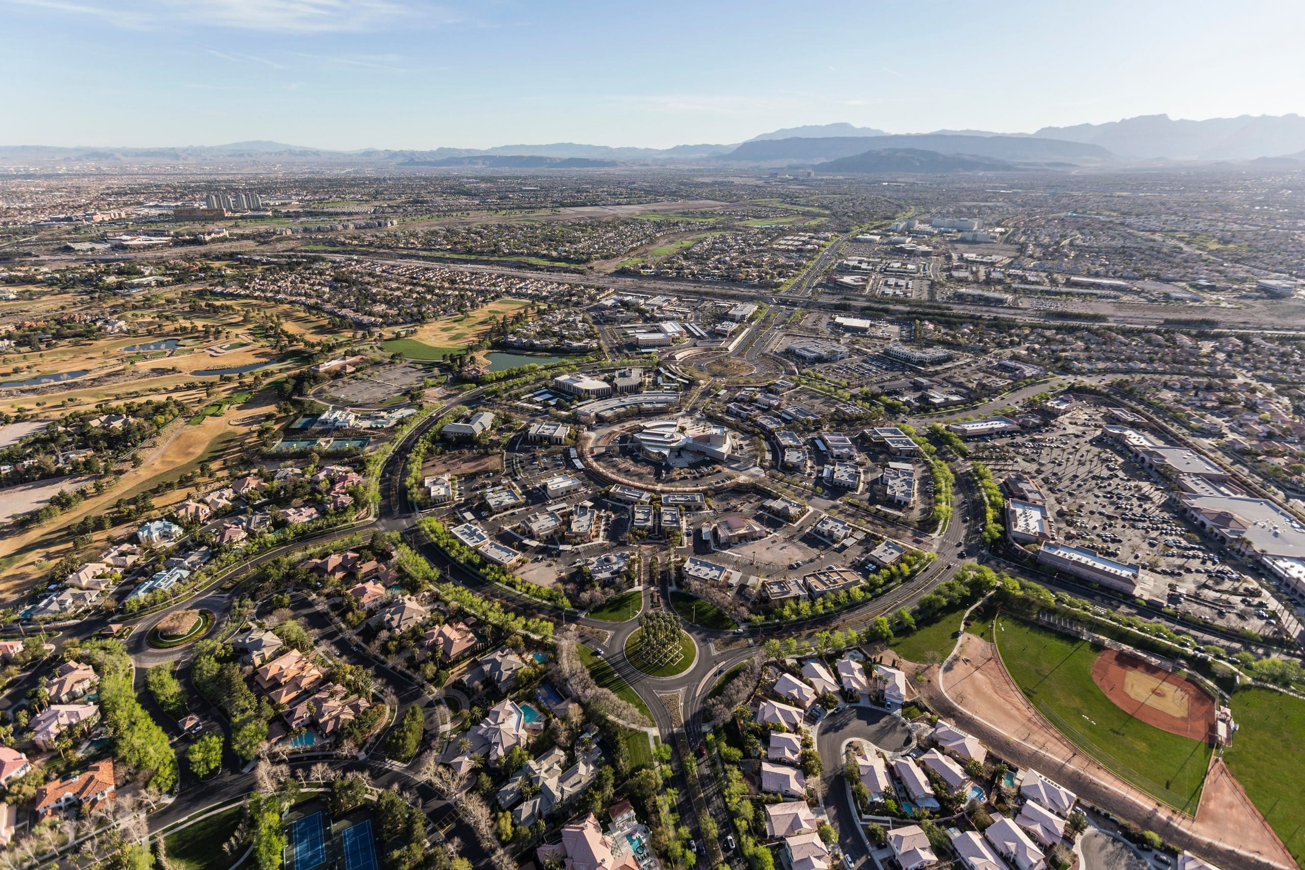 An aerial view of a suburban area of of homes in Summerlin NV, showcasing a mix of residential neighborhoods and commercial centers. The layout features a circular central area, possibly a shopping or business district, surrounded by lush green spaces, including a golf course. The backdrop includes distant mountains, which add a scenic contrast to the urban landscape below. The design of the community, with its curving roads and well-maintained properties, suggests a planned urban environment. This type of setting is typical of modern suburban developments found in regions that balance urbanization with accessible outdoor amenities.