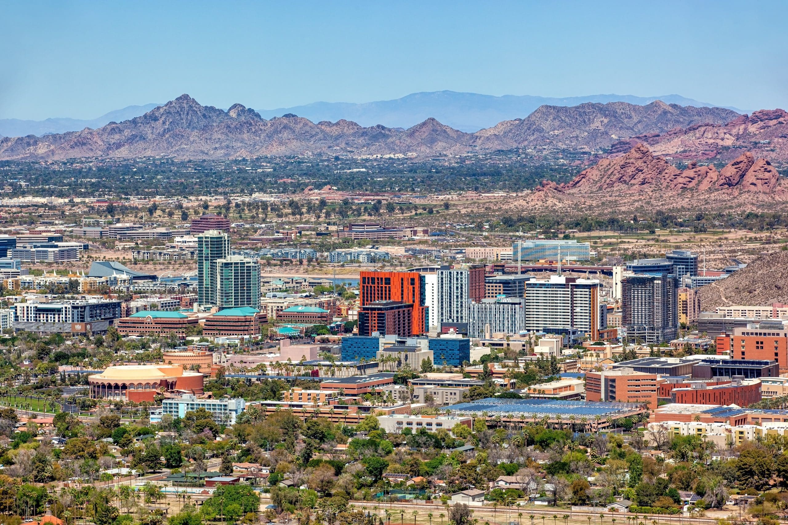 A panoramic view of a modern cityscape in Tempe AZ with an impressive backdrop of rugged mountains. The city is dense with various high-rise buildings featuring contemporary architectural styles. The predominant colors of the buildings range from neutral tones to striking reds and blues. There are also some green spaces scattered throughout the city. In the distance, the mountain range adds a dramatic natural element to the urban environment, highlighting the unique geographic setting of this location.