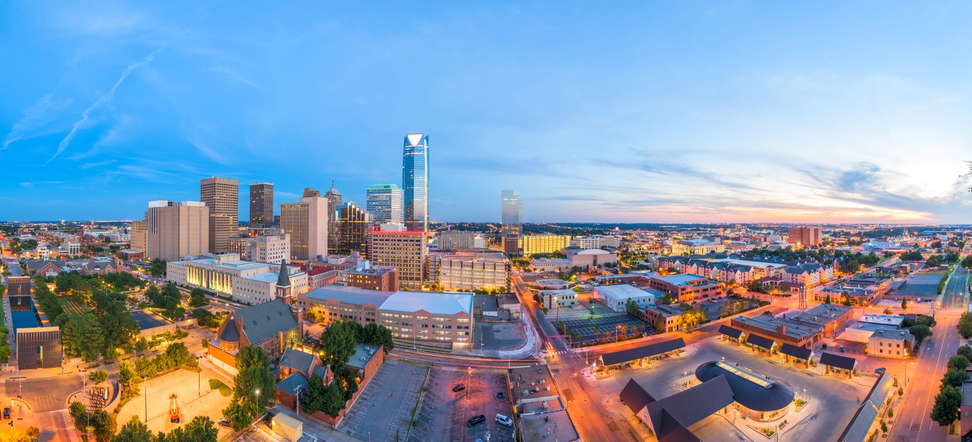 A panoramic view of downtown Oklahoma City, Oklahoma, showcasing its skyline at sunset. The photo highlights prominent buildings, green spaces, and urban architecture, giving a vibrant and warm aesthetic.